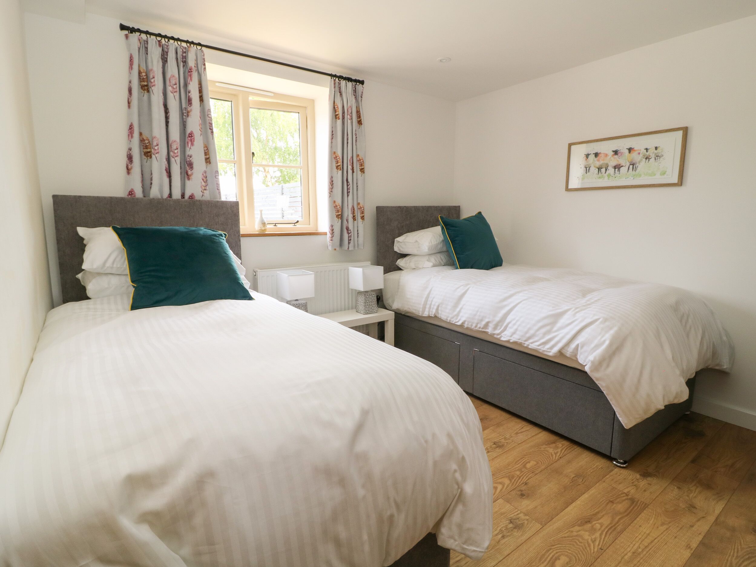 A hotel room with two beds, a window with curtains, a bedside table with a lamp, and a framed picture on the wall.