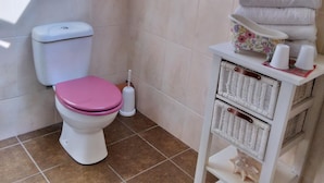 Un baño con tragaluz, inodoro con asiento rosa, escobilla de inodoro blanca y estante con cestas y toallas.