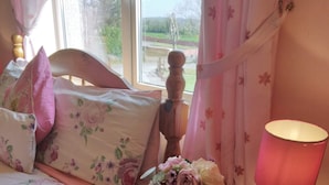Un dormitorio con cama, una mesita de noche con lámpara, un florero con flores y una ventana con cortinas.