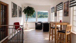 A modern interior with a wooden floor, a dining table with chairs, a red chair, a black chair, a potted plant, and a view of a lake.
