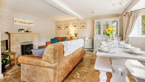 A cozy living room with a fireplace, a sofa, a dining table set for a meal, and a kitchen area with cabinets and a window. - Traditional Cotswold stone cottage sleeps 7 guests in 3 bedrooms - Little Mullions (Little Rissington, near Bourton-on-the-Water)