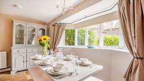 A dining area with a white table set for four, a cabinet with glass doors, and a window with curtains. - Traditional Cotswold stone cottage sleeps 7 guests in 3 bedrooms - Little Mullions (Little Rissington, near Bourton-on-the-Water)