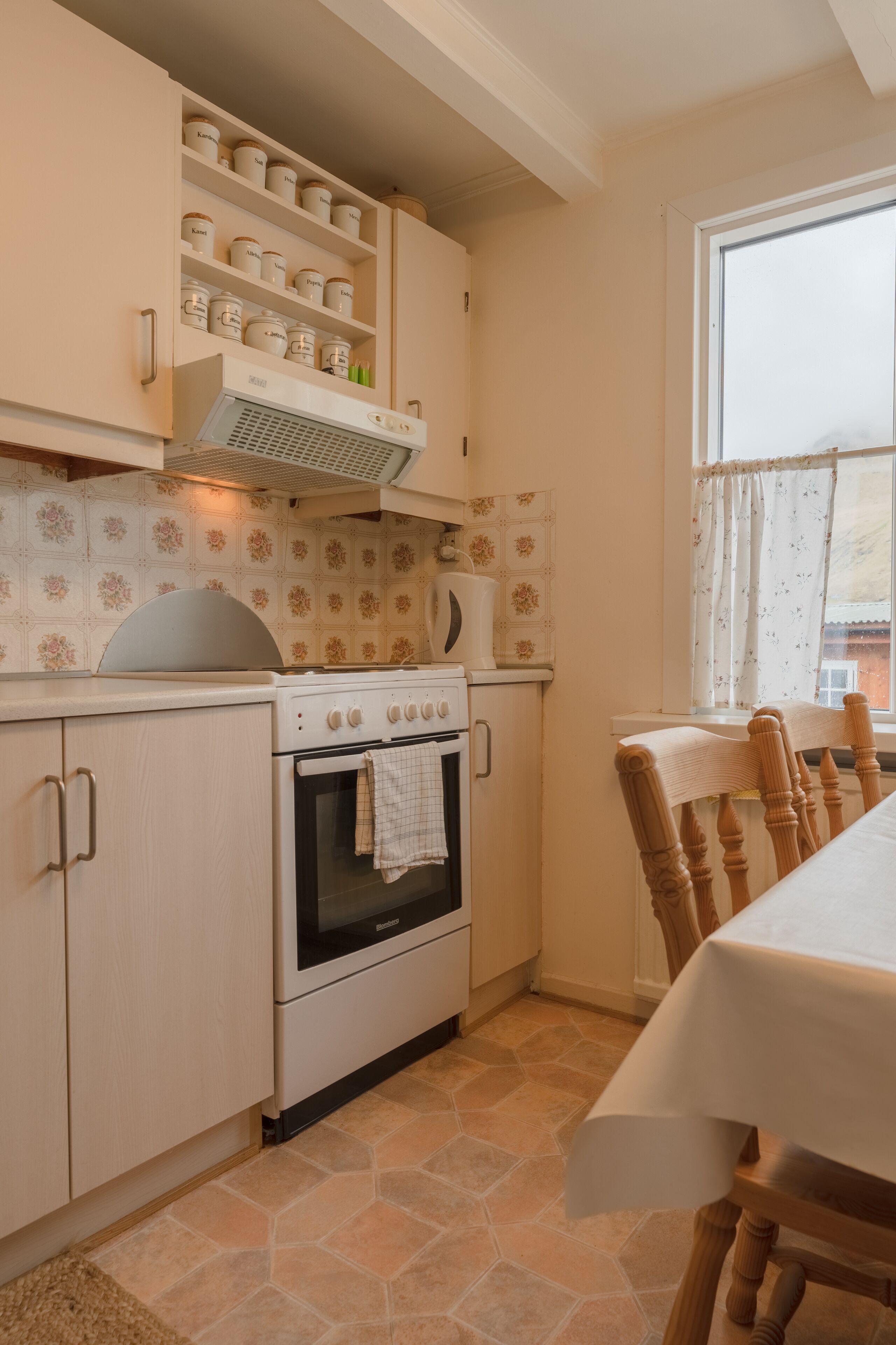 Una cocina con estufa, horno y alacenas. Hay una ventana con cortinas y una mesa con sillas.
