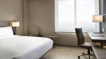 A hotel room with a large bed, a desk, a chair, a small table, and a window with a city view.