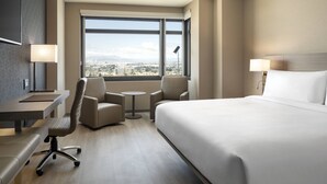 Hotelkamer met een groot bed, een bureau, twee stoelen, een kleine tafel en een stadszicht door grote ramen.