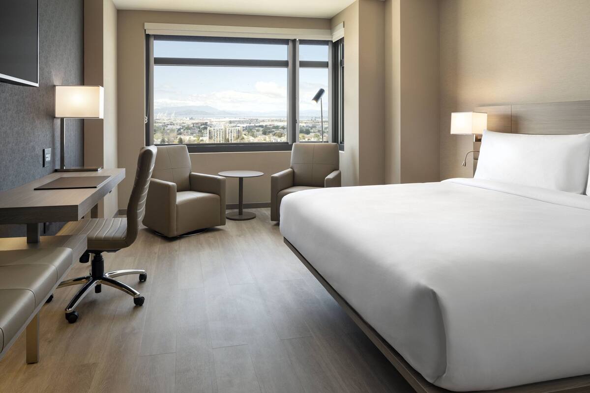 A hotel room with a large bed, a desk, two chairs, a small table, and a view of a cityscape through large windows.