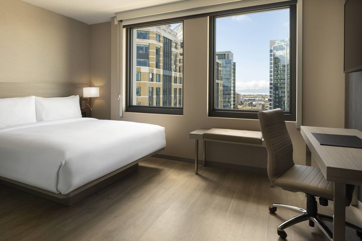 A hotel room with a bed, desk, chair, and large windows offering a city view.