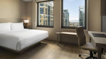 A hotel room with a bed, desk, chair, and large windows offering a city view.