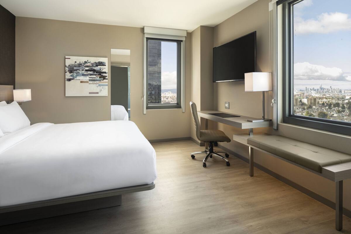 A hotel room with a bed, desk, chair, and a large window offering a city view.