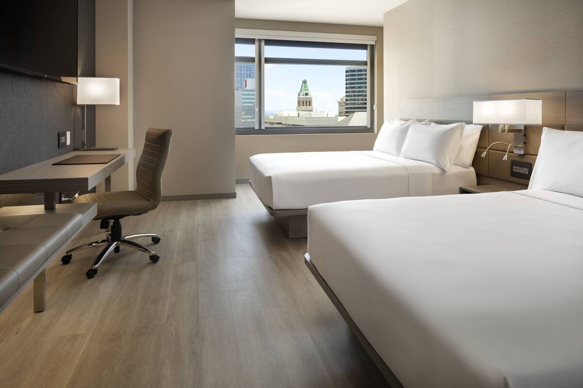 A hotel room with two beds, a desk, a chair, and a large window with a city view.