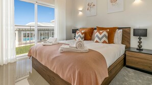 A hotel room with a large bed, a nightstand, a lamp, and a view of a pool and building through a window. - Casa Makin at Boa Vista Golf Resort, Bayview Village (Lagos)