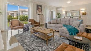 A modern living room with a large sofa, a coffee table, and a dining area. There is a large window providing a view of the outdoors. - Casa Makin at Boa Vista Golf Resort, Bayview Village (Lagos)