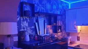 Ein Zimmer mit Küchenzeile, ausgestattet mit einem weißen Schrank, einer Arbeitsplatte und einem dekorativen Spiegel. Regale mit verschiedenen Gegenständen und eine Wand mit tropischem Muster.