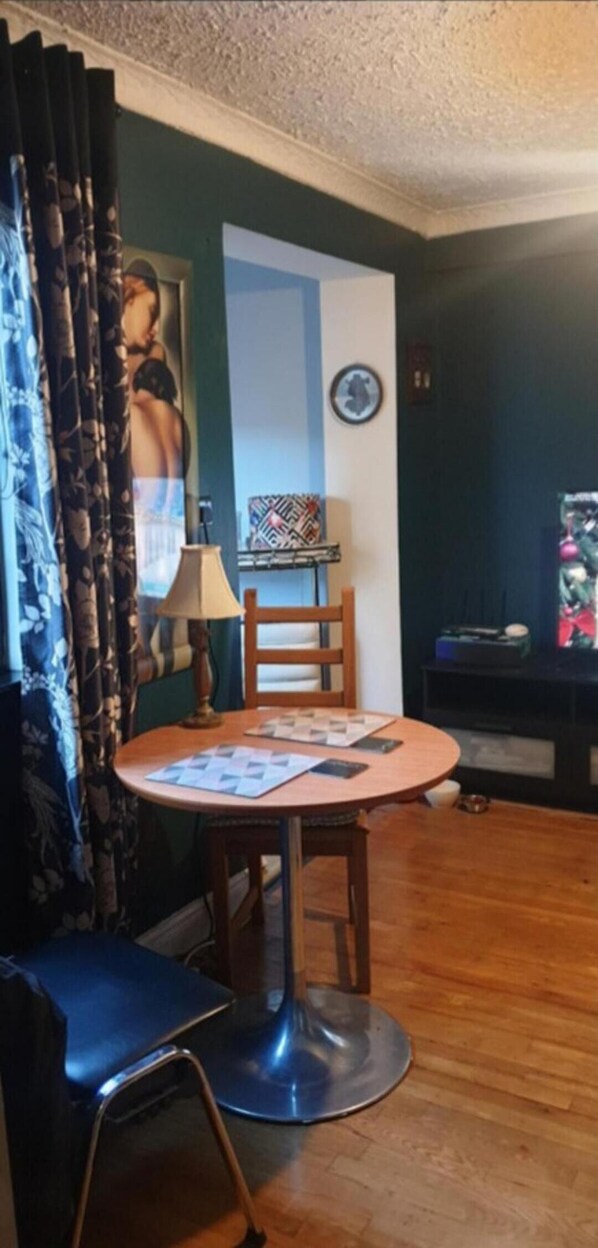 Um quarto pequeno e aconchegante com uma mesa de madeira redonda, uma cadeira azul, uma luminária e um cartaz na parede.