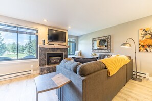 A modern living room with a fireplace, a sofa, a TV, and a large window offering a view of trees.