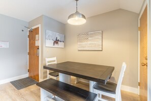 A dining area with a dark wooden table and white chairs, a pendant light, a wooden door, and a framed picture on the wall.