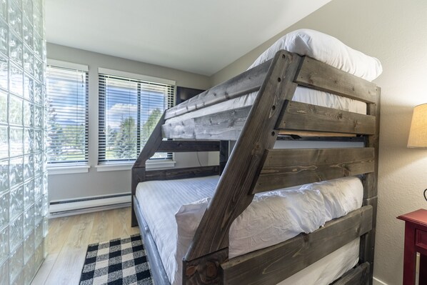 A bunk bed with a wooden frame and white bedding, a checkered rug, and a window with blinds.