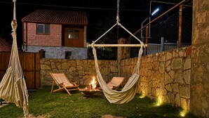Un fauteuil suspendu est installé au-dessus d’un foyer dans un jardin, avec un mur de pierres et une maison en arrière-plan.