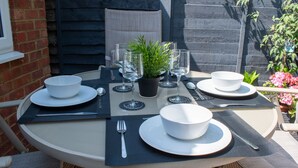 A round table set for four with white plates, bowls, cutlery, and clear wine glasses, accompanied by a potted plant. - 121 Studio (Bishop's Stortford)