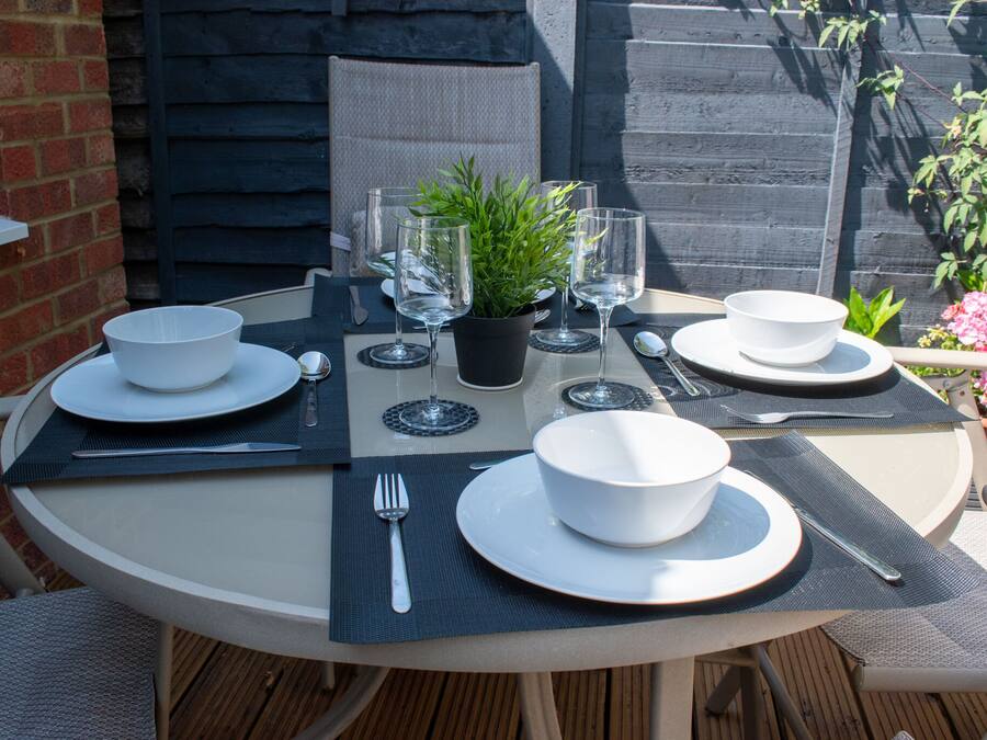 A round table set for four with white plates, bowls, cutlery, and clear wine glasses, accompanied by a potted plant.