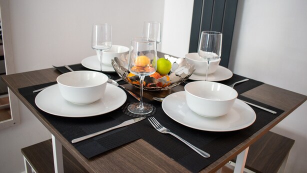 A dining table set with white bowls, plates, cutlery, and a fruit bowl with a glass of water. - 121 Studio (Bishop's Stortford)