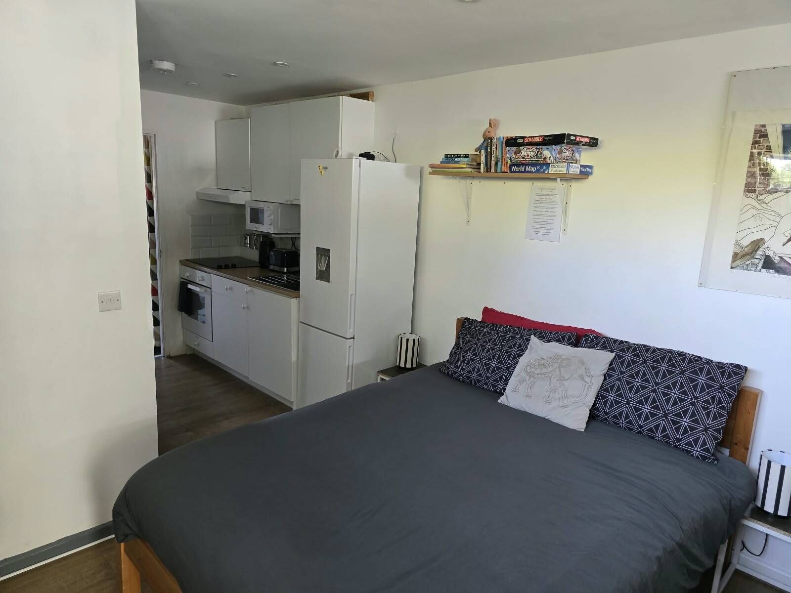 Een compacte slaapkamer met een bed, een kleine kitchenette en een plank met boeken en spelletjes.