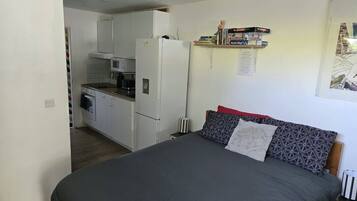 Een compacte slaapkamer met een bed, een kleine kitchenette en een plank met boeken en spelletjes.