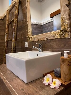 A bathroom with a white sink, a wooden vanity, a mirror with ornate woodwork, and a wooden soap dispenser. - Mikuk Cottages Canggu (Canggu)