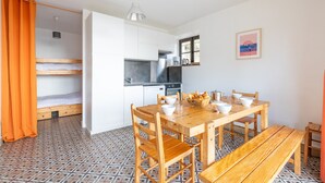 A compact room with a wooden dining table, benches, and a kitchen area with white cabinets and a black stove. - Garden level with sea view in the heart of Pléneuf (Pléneuf-Val-André)