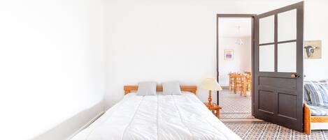 A single bed with a wooden frame and white bedding, a small bedside table with a lamp, a door leading to a dining area, and a patterned floor.