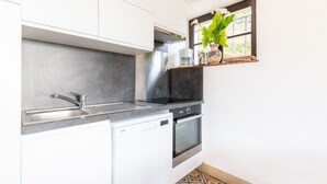 A compact kitchen with white cabinets, a built-in dishwasher, a stove, and a window with a plant. - Garden level with sea view in the heart of Pléneuf (Pléneuf-Val-André)