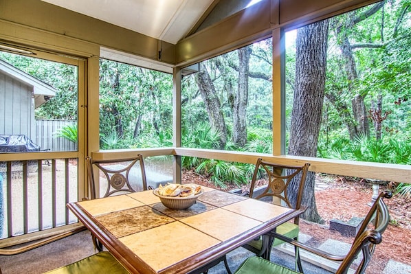 Uma área de refeições ao ar livre coberta, com mesa e cadeiras, rodeada por amplas janelas com vista para uma área arborizada.