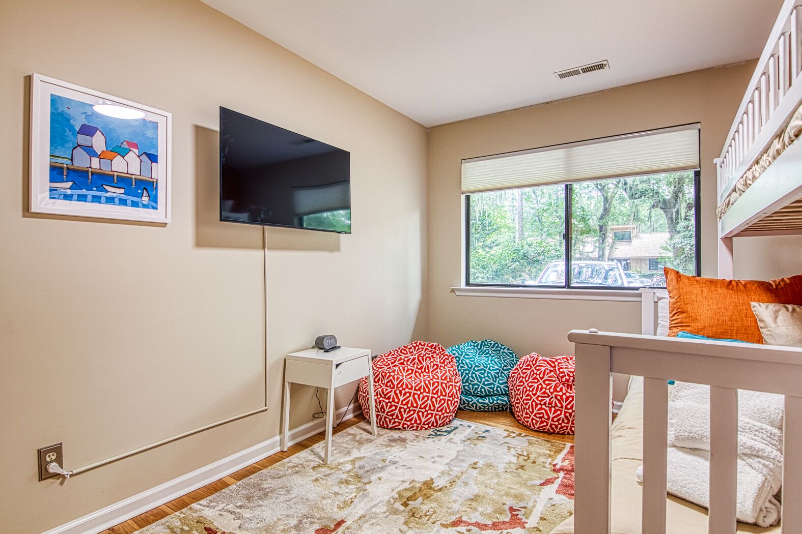 A child's room with a bunk bed, a flat-screen TV, a small bedside table, a window with a view of greenery, and colorful bean bags.