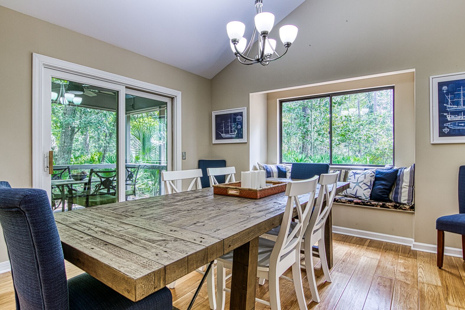 Un espace repas avec une table en bois, des chaises blanches et un banc agrémenté de coussins. De grandes fenêtres offrent une vue sur la verdure.
