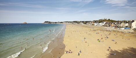 A wide sandy beach with clear blue waters, a rocky shoreline, and buildings along the coast.