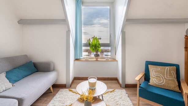 A modern living room with a grey sofa, a blue armchair, and a small round table with a glass of milk. A window with a view of the sky and a plant on the windowsill is visible. - 50m from the beach, charming flat with sea view (Pléneuf-Val-André)