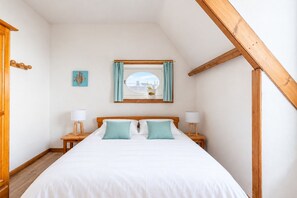 A bedroom with a bed, two bedside tables, a window with curtains, a wall-mounted picture, and a wooden ceiling beam. - 50m from the beach, charming flat with sea view (Pléneuf-Val-André)