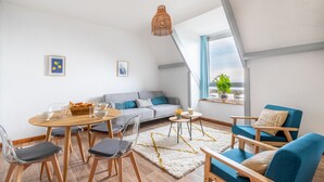 A modern living room with a dining area, grey sofa, blue armchairs, wooden flooring, and a large window with a view. - 50m from the beach, charming flat with sea view (Pléneuf-Val-André)