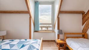 A bedroom with a sloped ceiling, a window with a curtain, a bed with a blue and white quilt, a bedside table with a lamp, and a wooden headboard. - 50m from the beach, charming flat with sea view (Pléneuf-Val-André)