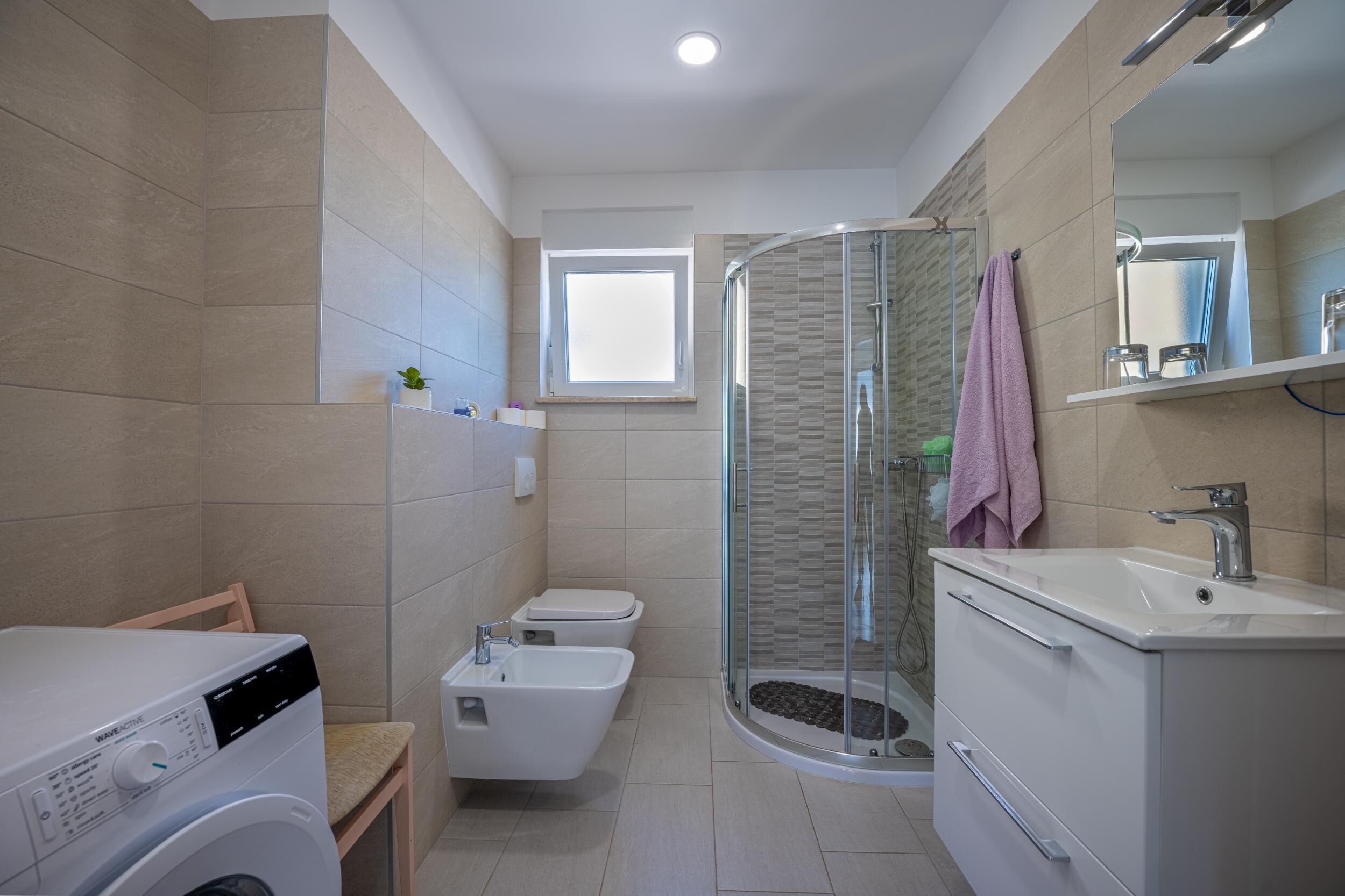 Een moderne badkamer met wasmachine, bidet, douchecabine van glas en een wastafel met spiegel.