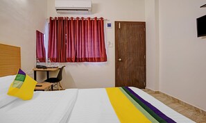 Une chambre d’hôtel avec un lit, un bureau et une chaise, un climatiseur, une porte en bois et une fenêtre avec des rideaux rouges.