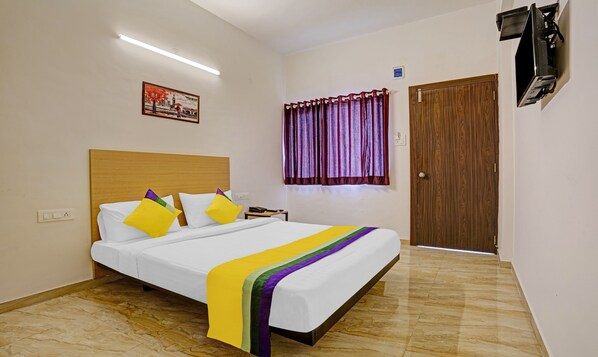 Une chambre d’hôtel comprenant un lit, une table de chevet, un téléphone, une fenêtre avec des rideaux, une porte et un téléviseur à écran plat fixé au mur.
