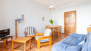 A living room with a wooden floor, a blue sofa, a striped armchair, a wooden dining table with chairs, a television, and a hanging light fixture. - Breathtaking view of the sea for 7 people in Pléneuf (Pléneuf-Val-André)