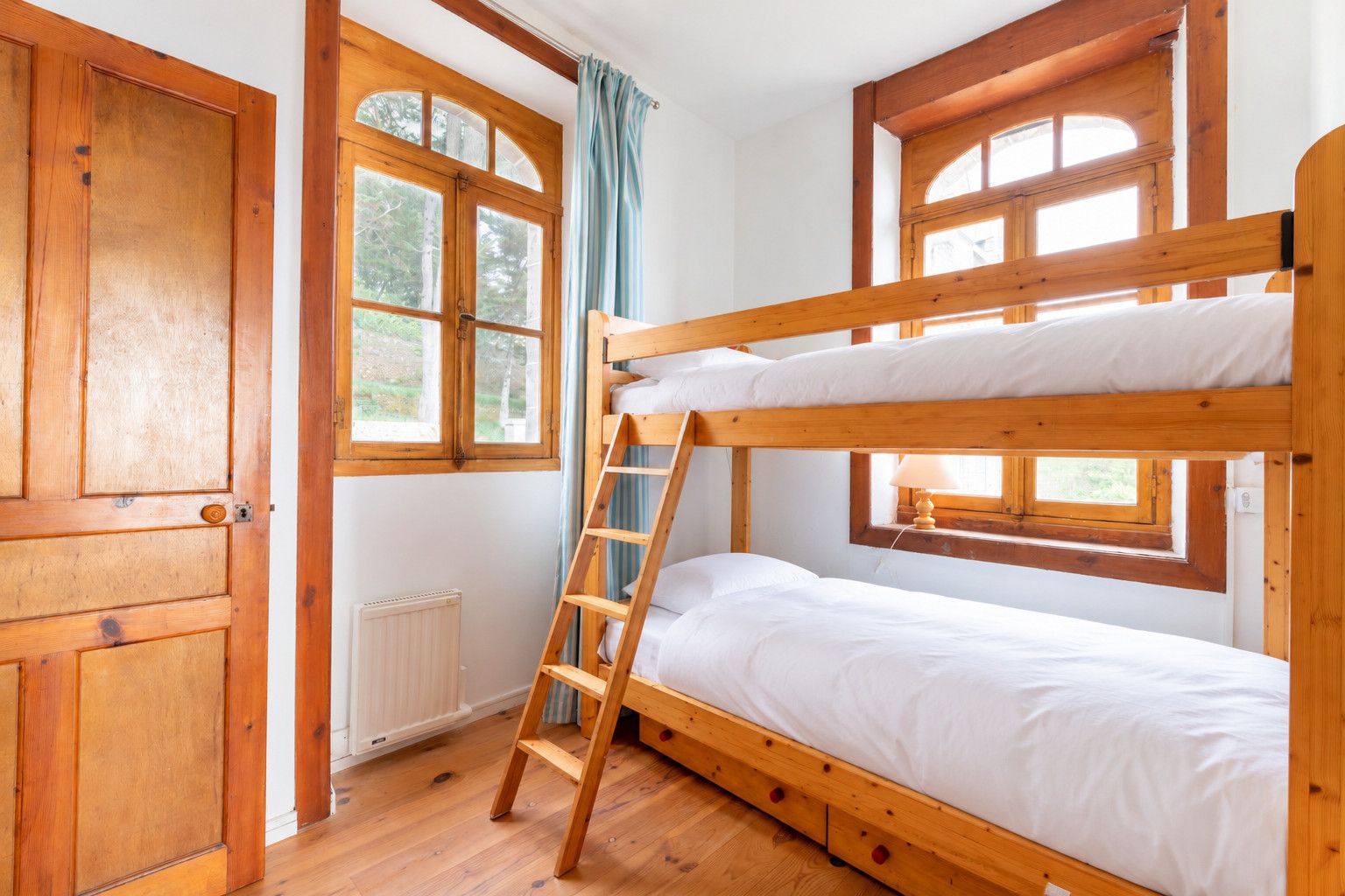 Une chambre avec un lit superposé, des poutres en bois, une échelle et une fenêtre avec des rideaux.
