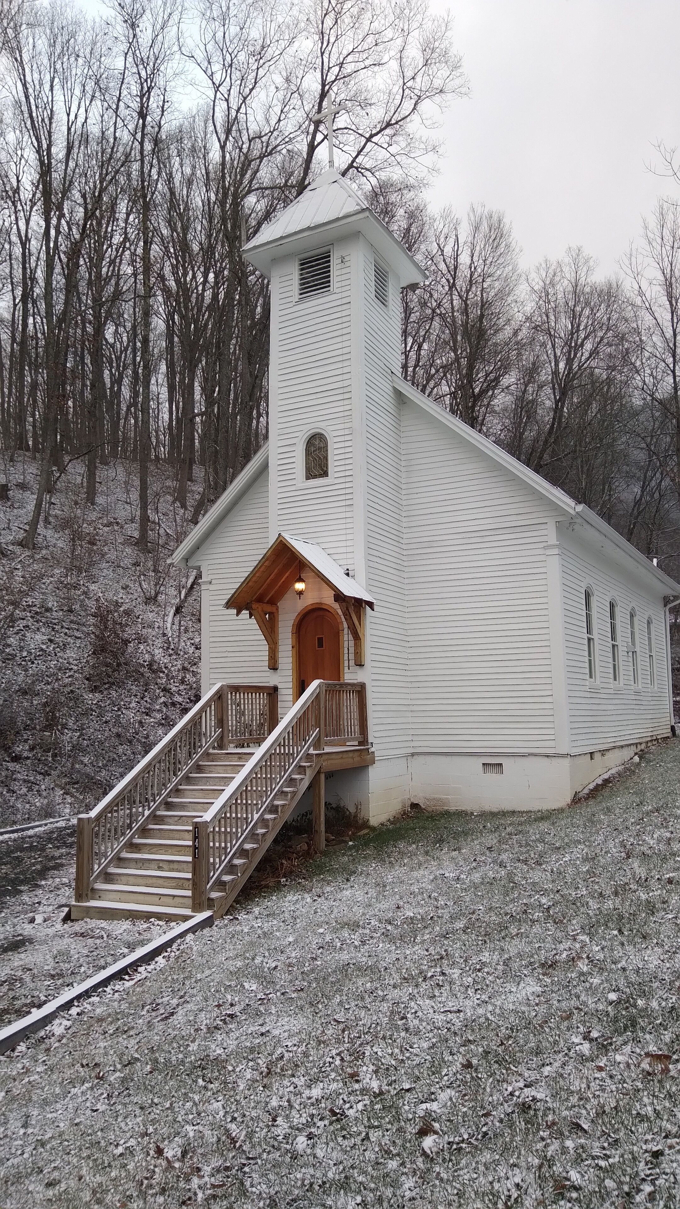 Une église blanche avec un escalier en bois menant à l’entrée, entourée d’un paysage enneigé et d’arbres.