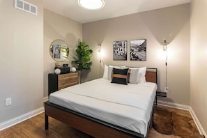 Un dormitorio con cama, mesitas de noche, un espejo, una obra de arte en la pared y una planta en maceta.