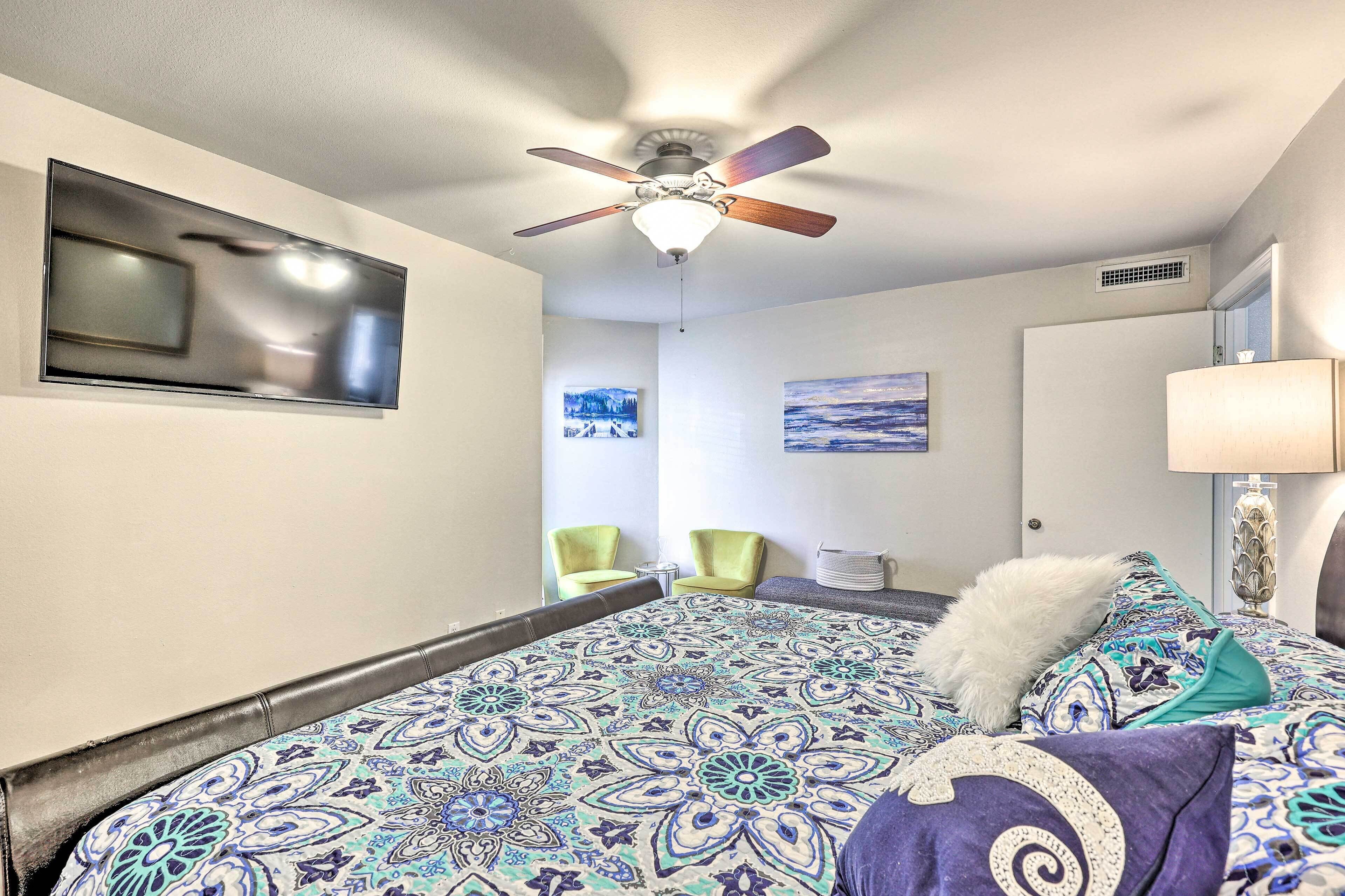 A bedroom with a bed, ceiling fan, wall-mounted TV, two chairs, and a lamp.