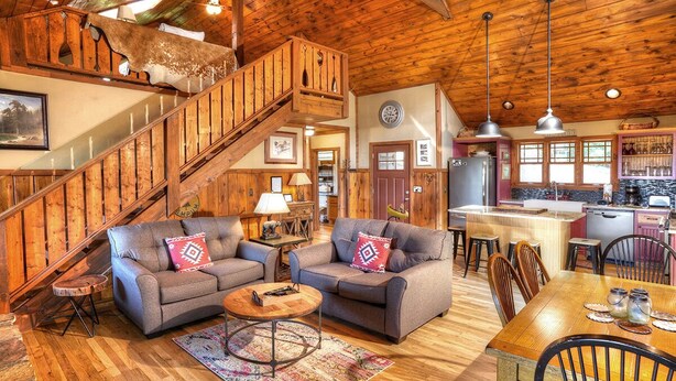 A rustic interior with a wooden staircase, two sofas with decorative pillows, a wooden dining table, and a kitchen area.
