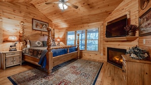 A wooden cabin bedroom with a bed, a fireplace, a television, and a window with blinds.