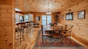 A wooden cabin interior with a dining area, bar stools, a large table, and a rug.